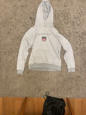 Grå hoodie från GANT med logga - Snygg grå hoodie från GANT med broderad logga och sköld på bröstet. Klassisk känguruficka framtill och mjuk huva. Perfekt för en avslappnad och stilren look.
