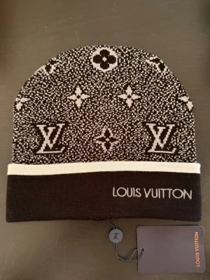 L mössa - Snygg svart mössa med klassiskt monogrammönster i vitt och kontrasterande vit kant. Perfekt accessoar för dig som vill ha en exklusiv och trendig look. Helt ny, oanvänd.