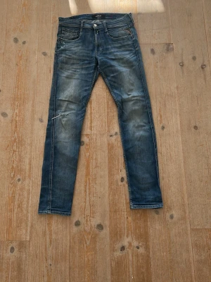 Blå slitna jeans från Replay - Säljer ett par blå jeans från Replay med snyggt slitna detaljer och en liten reva på vänster lår. Jeansen har klassisk femficksmodell, raka ben och kontrastsömmar. Perfekta för en avslappnad och trendig look.