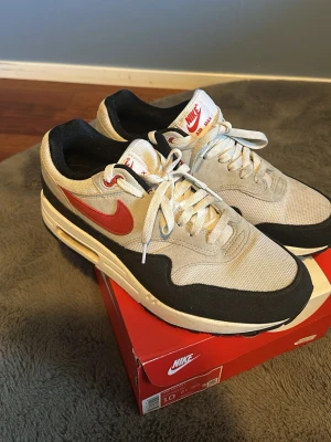 Nike Air Max 1 - Nike Air Max 1 sneakers i vitt mesh och mocka med svarta och grå detaljer samt röd Swoosh. Klassisk siluett med synlig Air-enhet i sulan och vita skosnören. Perfekta för dig som gillar retrostil och streetwear.