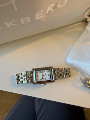 Silverfärgad klocka från Mockberg - Stilren klocka från Mockberg med rektangulärt silverfärgat boett och länkarmband i metall. Vit urtavla med romerska siffror och tunna svarta visare. Kommer med extra länkar och vit förvaringspåse.