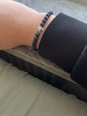 Svart lavastenarmband med silverdetalj - Snyggt armband med svarta lavastenpärlor och en cool silverfärgad detalj i mitten. Elastiskt band som gör det enkelt att ta på och av. Perfekt accessoar för dig som gillar stilrena och trendiga smycken.