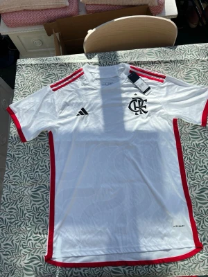 Vit Flamengo matchtröja Adidas - Snygg vit matchtröja från Adidas med Flamengo-logga på bröstet, röda detaljer längs sidor och axlar samt svarta Adidas-ränder. Tillverkad i lätt och ventilerande material med Aeroready-teknologi. Perfekt för fotboll eller träning.