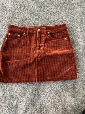 Rostbrun manchesterkjol med fickor - Säljer en snygg kortkjol i rostbrun manchester med klassiska fickor fram och bak samt bälteshällor. Kjolen har en rak passform och stängs med knapp och dragkedja framtill. Perfekt för dig som gillar retrovibbar och mjuka material.