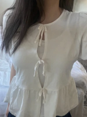 Vit blus med knyt och volang - Söt vit blus med tre dekorativa knyt framtill och volang nedtill. Blusen har rund halsringning och korta puffärmar som ger en romantisk vibe. Tillverkad i ett lätt tyg som känns luftigt och skönt. Perfekt att styla med jeans eller kjol.