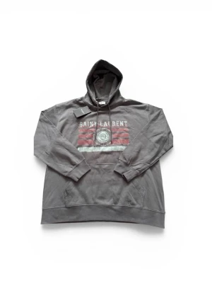 Grå Saint Laurent hoodie med tryck - Säljer en grå hoodie från Saint Laurent med stor huva och känguruficka. På bröstet finns ett stort tryck med texten 'SAINT LAURENT' och röda samt vita ränder, samt ett SL-emblem i mitten. Hoodien har ribbade muddar och är tillverkad i mjuk bomull.