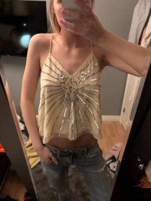 Beige paljettlinnetopp med smala band - Säljer en beige linnetopp med tunna axelband och v-ringning. Toppen har ett glittrigt mönster av silverpaljetter. Passar någon som har S 