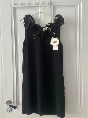 Svart klänning med volangdetaljer - Twist & Tango - Säljer en svart ärmlös klänning från Twist & Tango med volangdetaljer runt halsringningen. Klänningen har en rak passform och är tillverkad i ett mjukt, glansigt material som ger en elegant look. Perfekt för dig som vill ha något stilrent och trendigt. Klänning har en inbyggd underklänning och fickor. Ej använd.