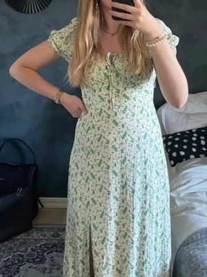 Grönblommig långklänning med puffärm - Långklänning i ljusgrönt och vitt med blommigt mönster. Klänningen har puffärmar, slits framtill och söt knytning vid bysten. Smockad rygg för skön passform och lätt volang i kjolen. Perfekt för sommarens alla tillfällen. Orginalpris 700