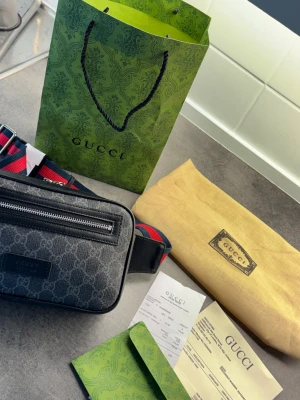 Gucci axelväska med GG-mönster - Snygg axelväska från Gucci i svart och grått GG-mönstrat canvasmaterial med detaljer i svart läder. Väskan har en justerbar axelrem i rött och blått, samt en praktisk ytterficka med dragkedja. Perfekt för dig som vill ha en trendig och lyxig accessoar.