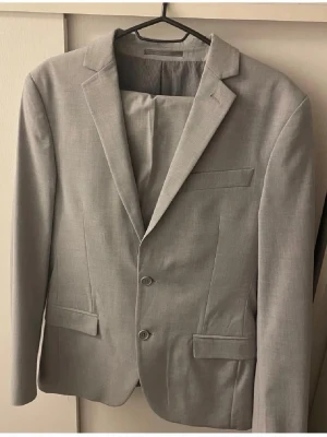 Ljusgrå klassisk kostym  - Snygg ljusgrå kostym med två knappar framtill, klassisk passform och smala slag. Kavajen har två fickor med lock och en bröstficka. Materialet ser ut att vara en tunnare vävd textil, perfekt för en stilren look.