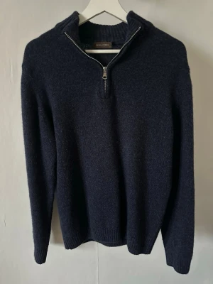 Mörkblå stickad halvzip-tröja - Mörkblå stickad tröja från Oscar Jacobson med halvzip och hög krage. Tröjan har långa ärmar, ribbstickade muddar och är tillverkad i ett mjukt, ulligt material. Enkel och stilren design som passar perfekt till kalla dagar.