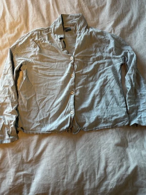 Randig pyjamaströja från Kappahl XS - Ljusblå och vitrandig pyjamaströja från Kappahl i storlek XS. Skjortmodell med långa ärmar, knäppning framtill och bröstficka. Tillverkad i mjukt och tunt tyg, perfekt för mysiga kvällar hemma.