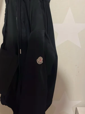 Svart vindjacka från Moncler - Svart vindjacka från Moncler med dragkedja framtill, huva med dragsko och Moncler-logga på ärmen. Jackan har en stilren design och tunnt material som passar perfekt för blåsiga dagar. Fickor med dragkedja och diskreta detaljer.