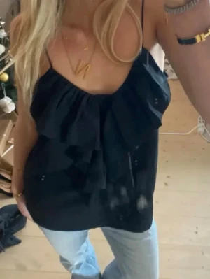 Svart volang topp - Snygg svart topp med tunna axelband och volangdetaljer framtill. Perfekt till jeans eller kjol för en trendig look. Luftigt material som känns skönt ger en avslappnad men chic vibe