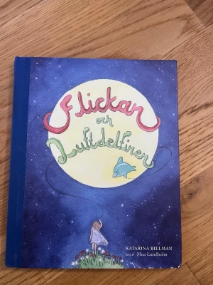 Flickan och Luftdelfinen bok - En fantasifull skönlitterär bok med hårda pärmar, blått ryggband och ett färgstarkt omslag med en flicka, en ballong och en delfin mot en stjärnhimmel. Skriven av Katarina Billman och illustrerad av Moa Lundholm.