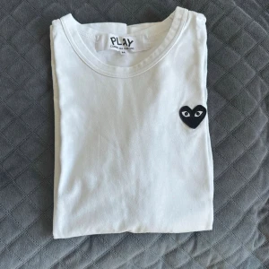 Vit t-shirt Comme des Garçons Play - Vit t-shirt från Comme des Garçons Play med det ikoniska svarta hjärta- och ögonmärket på bröstet. Klassisk rund hals och enkel design som passar till allt. Tillverkad i mjuk bomull för skön känsla. Perfekt inför sommaren och väldigt bra skick. Skriv om ni vill ha mer info eller bilder. Kan inte visa passfor på grund av att den är för liten för mig!!