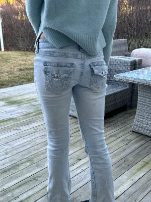 Ljusblå bootcut jeans True Religion - Säljer ett par ljusblå bootcut jeans från True Religion med snygga detaljerade bakfickor och låg midja. Jeansen har klassisk femficksdesign, knappgylf och är tillverkade i mjuk denim med lätt tvättad look.