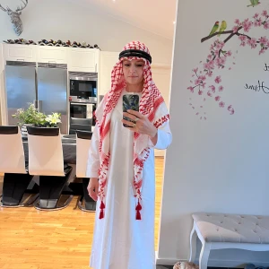 Arabisk shejk maskeradklädsel - Maskeradklädsel med lång vit kaftan, rödvit mönstrad sjal med tofsar och svart huvudband. Perfekt för temafester eller cosplay. Materialet är tunt tyg, troligen polyester eller bomullsblandning. Klassisk look inspirerad av traditionell arabisk shejk-stil. Köpte i Dubai till min pojke som var 13 år då men den passar även mig som har storlek S