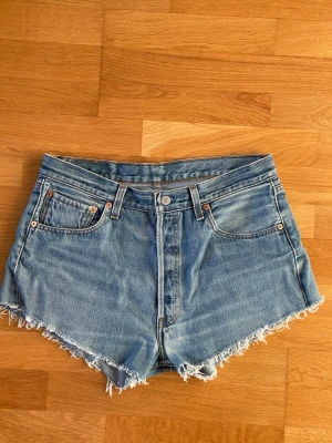 Levi's 501 ljusblå jeansshorts med fransar storlek W31 - Säljer ett par klassiska ljusblå jeansshorts från Levi's 501 med fransig rå kant och femficksmodell. Shortsen har hög midja och är tillverkade i slitstark bomull. Perfekta för sommaren och har den ikoniska Levi's-lappen bak. Storlek W31