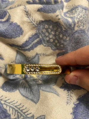 Guldigt armband med gnistrande pärlor - Snyggt armband i guldfärgad metall med glittrande små pärlor och stenar på ovansidan. Armbandet har en modern, stilren design med en rektangulär detalj fylld av gnistrande dekorationer. Perfekt för dig som vill ha något som sticker ut.