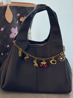  handväska med charms - Trendig brun/röd handväska i mjukt konstläder. Endast använd några få gånger. Otroligt fin kvalite och har 3 fack invändigt. Väsksmycket ingår samt ett långt band. (På bild 3 är smutsen på spegeln inte på väskan😅)