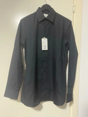 Svart slim fit skjorta från Next - Stilren svart skjorta från Next Tailoring med klassisk krage och slim fit passform. Skjortan har långa ärmar och knappar framtill. Tillverkad i bomull för en bekväm känsla och snygg look. Perfekt för dig som gillar en enkel men elegant stil.