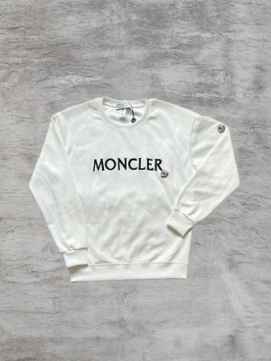Vit sweatshirt från Moncler - Stilren vit sweatshirt från Moncler med svart logotyptryck på bröstet och en liten Moncler-patch på ärmen. Tröjan har rund halsringning, ribbade muddar och är tillverkad i mjukt material. Perfekt för en clean och trendig look.