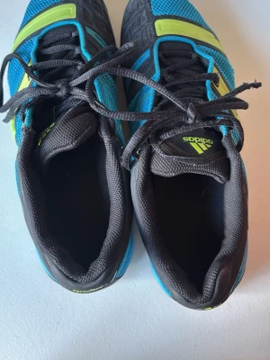 Blå Adidas sneakers med gula detaljer - Säljer ett par snygga Adidas sneakers i blå mesh med svarta och limegula detaljer. Skorna har klassisk snörning, svart vadderad plös och insida samt märkning på plösen och sidan. Perfekta för dig som gillar sportig stil och färg.