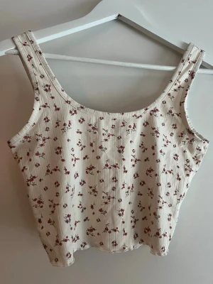 Blommig ribbad linnetopp i vitt - Söt ribbad linnetopp i vit nyans med små blommor i rött och rosa. Toppen har breda axelband och en rak, något croppad passform. Perfekt för sommardagar och enkel att matcha med jeans eller kjol.