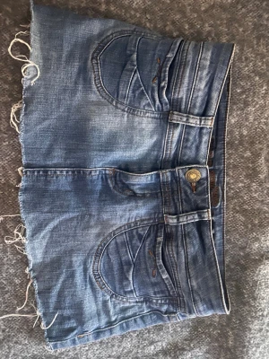 Blå jeanskjol med rå kant - Säljer en blå jeanskjol med rå, fransig kant och klassiska fickor både fram och bak. Kjolen har bälteshällor och stängs med knapp och dragkedja. Perfekt för dig som gillar en avslappnad och trendig stil.