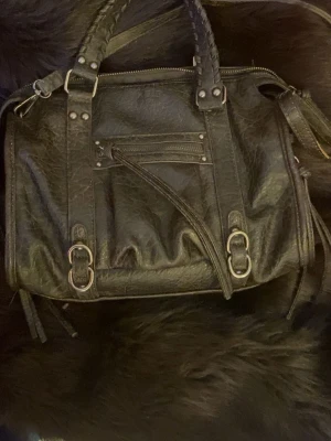 City bag - Snygg svart handväska från Fast med flätade handtag och detaljer i silverfärgad metall. Väskan har dragkedja framtill, dekorativa spännen och flera remmar. Tillverkad i skinnimitation med en cool, lite sliten look.