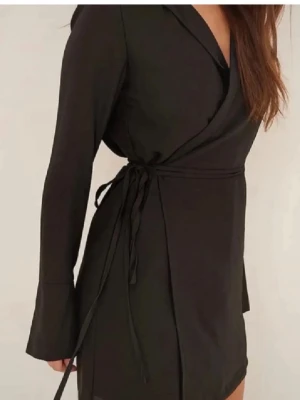Svart omlottklänning NA-KD - Snygg svart omlottklänning från NA-KD med långa ärmar och knytband i midjan. Klänningen har en kort längd och klassisk krage, perfekt för en trendig och stilren look.