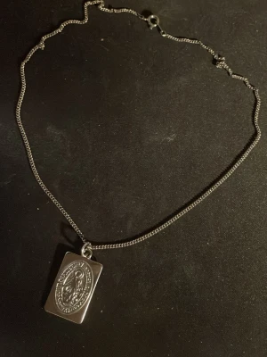 Silverfärgat halsband med rektangulärt hänge - Snyggt silverfärgat halsband med en tunn kedja och ett rektangulärt hänge med ingraverat motiv. Hänget har en oval detalj i mitten med ett mönster som ger en cool och unik look. Perfekt accessoar för att lyfta din outfit.OBS! Materialet är okänt, kan ej garantera att det är rostfritt.