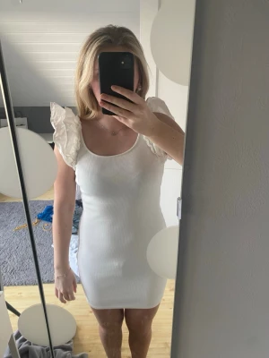 Vit ribbad kortklänning med volangärm - Säljer en vit ribbad kortklänning med bred rundringning och volangdetaljer på axlarna. Klänningen har en kroppsnära passform och är ärmlös med dekorativa volangärmar som ger en romantisk touch. Perfekt för sommarens alla tillfällen!