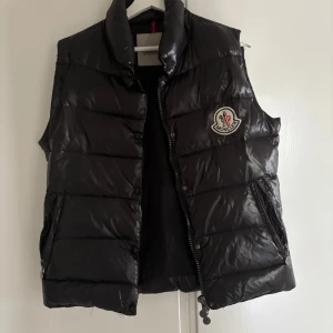 Svart dunväst från Moncler - Snygg svart dunväst från Moncler med dragkedja och broderad logga på bröstet. Perfekt för kyliga dagar när du vill hålla stilen. Västen har en quiltad design och praktiska fickor med dragkedja. Priset kan självklart diskuteras, använt ett fåtal gånger typ 2 och har inga minsta lilla fel.😁