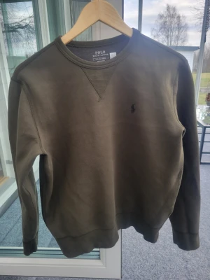 Olivgrön sweatshirt från Polo Ralph Lauren - Snygg olivgrön sweatshirt från Polo Ralph Lauren med rund halsringning och diskret broderad logga på bröstet. Tröjan har ribbade muddar vid ärmslut och nederkant, perfekt för en avslappnad och stilren look. Säljer denna pga att den blivit för liten. Nypris 1200 kr. 