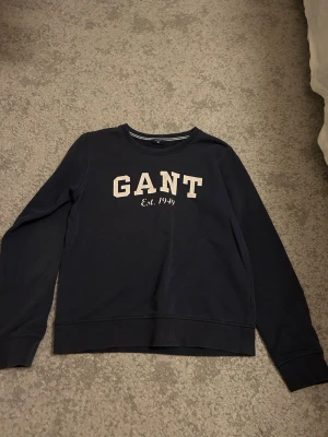 Mörkblå sweatshirt från GANT - Mörkblå sweatshirt från GANT med vit logga och texten 'Est. 1949' på bröstet. Tröjan har rund halsringning, långa ärmar och ribbade muddar vid ärmslut och nederkant. Perfekt för en avslappnad och stilren look.
