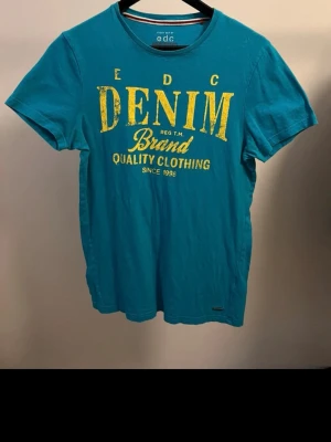 Blå t-shirt från EDC med tryck - Snygg blå t-shirt från EDC med stort gult tryck på bröstet där det står 'DENIM Brand Quality Clothing'. Klassisk rund halsringning och korta ärmar. Perfekt till jeans eller shorts för en avslappnad look.
