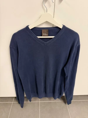 Blå V-ringad tröja Oscar Jacobson, stl L - Mörkblå stickad tröja från Oscar Jacobson med V-ringning och långa ärmar. Tröjan har en klassisk, stilren design och är tillverkad i ett mjukt, tunt material som passar perfekt till lager-på-lager. Ribbstickade muddar vid hals, ärmslut och nederkant.