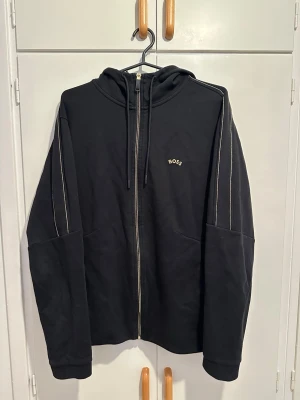 Svart zip hoodie från BOSS - Svart hoodie med dragkedja från BOSS. Hoodien har guldiga kontrastsömmar längs ärmar och huva, justerbar dragsko i huvan och en liten BOSS-logga på bröstet. Tillverkad i mjukt material, perfekt för en avslappnad och stilren look.