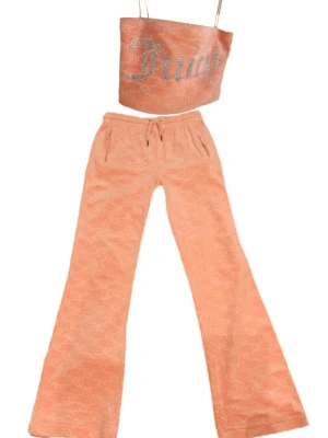 Juicy Couture peach loungewear set - Snyggt loungewear-set från Juicy Couture i persikofärgad velour. Setet består av ett linne/tubtopp med smala axelband och strassdetaljer samt matchande byxor med utsvängda ben och fickor. Både linne och byxor har Juicy-logga och JC-mönster över hela tyget. Byxornas fickor har dragkedjor.