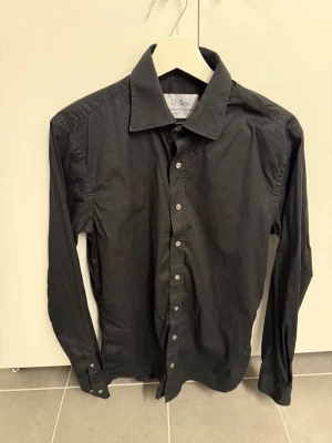 Svart skjorta från Oscar Jacobson, Stl 40 - Klassisk svart skjorta från Oscar Jacobson med lång ärm och knappar framtill. Skjortan har en stilren design med normal passform och är tillverkad i mjuk bomull. Perfekt till både jeans och kostymbyxor.