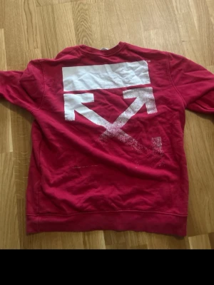 Röd sweatshirt från Off-White - Säljer en röd sweatshirt från Off-White med det ikoniska vita pilar-trycket på ryggen. Tröjan har rund halsringning och är tillverkad i mjukt bomullsmaterial. Perfekt för dig som gillar streetwear och vill sticka ut med en unik design.