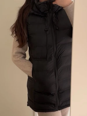 Svart lång dunväst med huva - Snygg svart dunväst med huva och dragsko, perfekt för lager på lager. Västen är quiltad och har fickor på sidorna samt dragkedja framtill. Passar till en avslappnad och trendig stil.