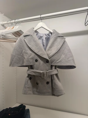Ljusgrå trenchcoat med cape - Stilren ljusgrå trenchcoat med dubbelknäppning, breda slag och bälte i midjan. Unik cape-detalj över axlarna ger en cool twist. Tillverkad i ett mjukt tyg som liknar ullmix. Perfekt för dig som vill sticka ut med en trendig och elegant look.