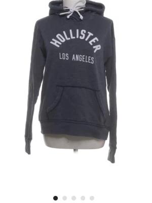 Mörkblå hoodie från Hollister - Mörkblå hoodie från Hollister med vit text 'Hollister Los Angeles' på bröstet. Klassisk känguruficka framtill, huva med vita dragsnören och ribbade muddar. Perfekt för en avslappnad och cool stil.