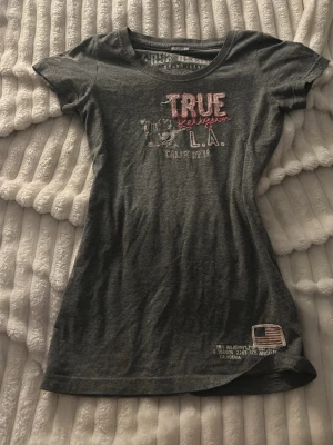 Mörkgrå True Religion t-shirt - Snygg mörkgrå t-shirt från True Religion med broderad text och patchdetalj framtill. T-shirten har rund halsringning och korta ärmar. Materialet är en mix av bomull och polyester, vilket gör den mjuk och bekväm. Diskuterar gärna priset.