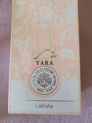 Yara Eau de Parfum från Lattafa - Yara Eau de Parfum från Lattafa kommer i en elegant ljusrosa kartong med blommigt mönster och gulddetaljer. Doften är en mix av fräscha och söta noter, perfekt för dig som gillar exklusiva och moderna parfymer. Flaskan rymmer 100 ml och har en lyxig känsla.