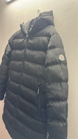Svart Moncler pufferjacka med huva - Svart pufferjacka från Moncler med huva och tydlig logga på ärmen. Jackan har dragkedja framtill, två sidofickor med dragkedja och stora Moncler-tryck på insidan. Materialet är glansigt och känns lyxigt, perfekt för kalla dagar.